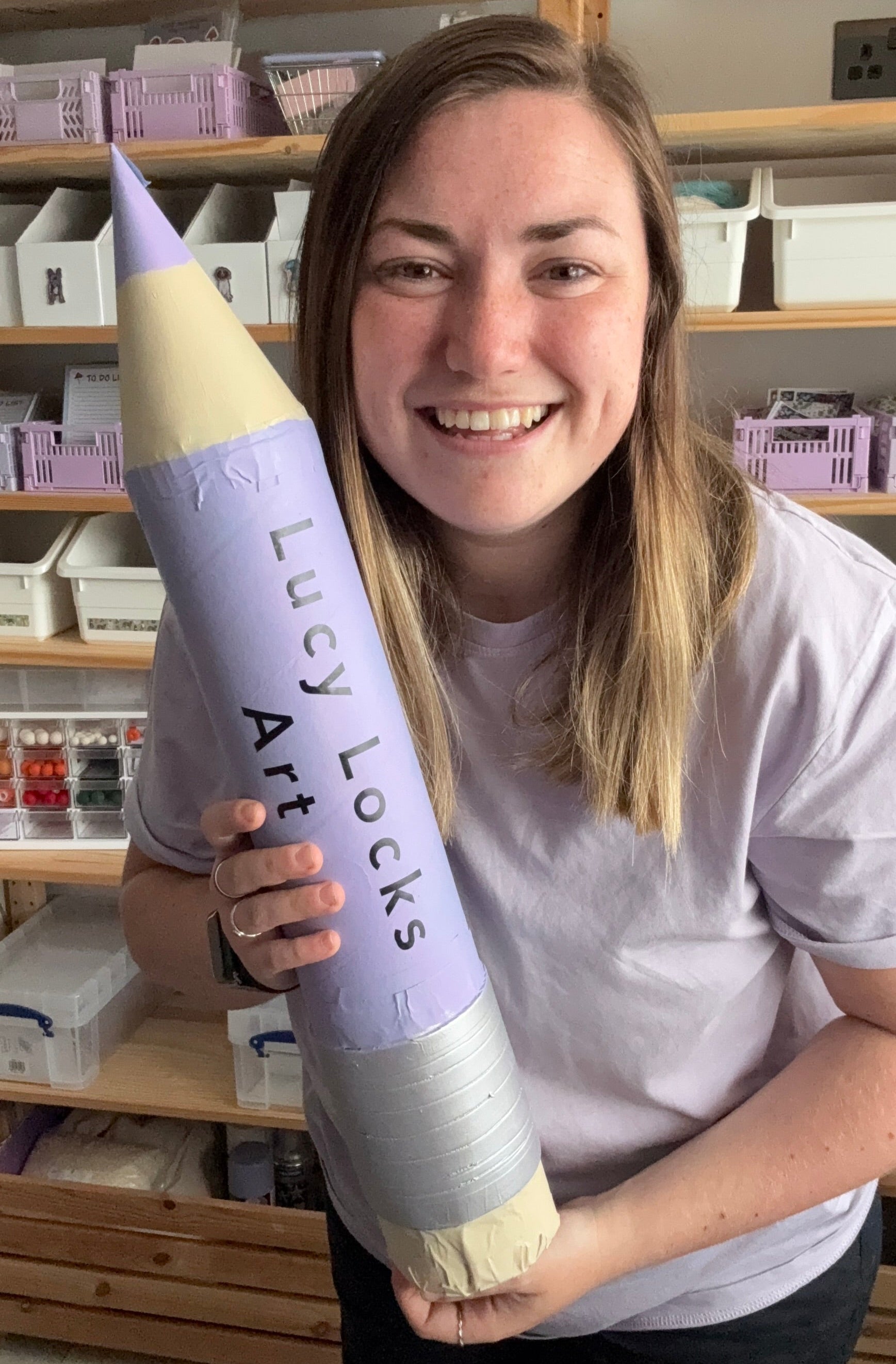 Lucy holding a large purple and yellow Pencil with 'Lucy Locks Art' branding in a store setting.