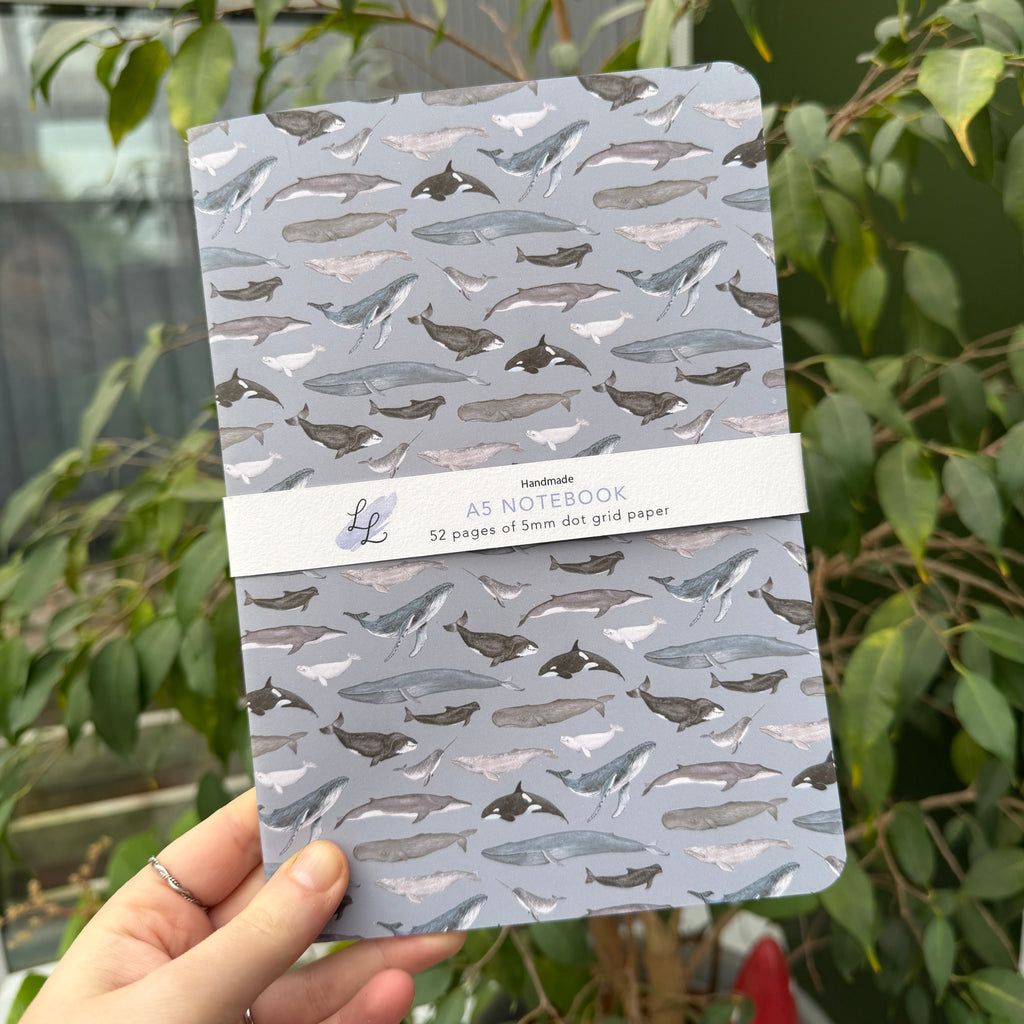 Hand holding a notebook with a whale design against a green outdoor background