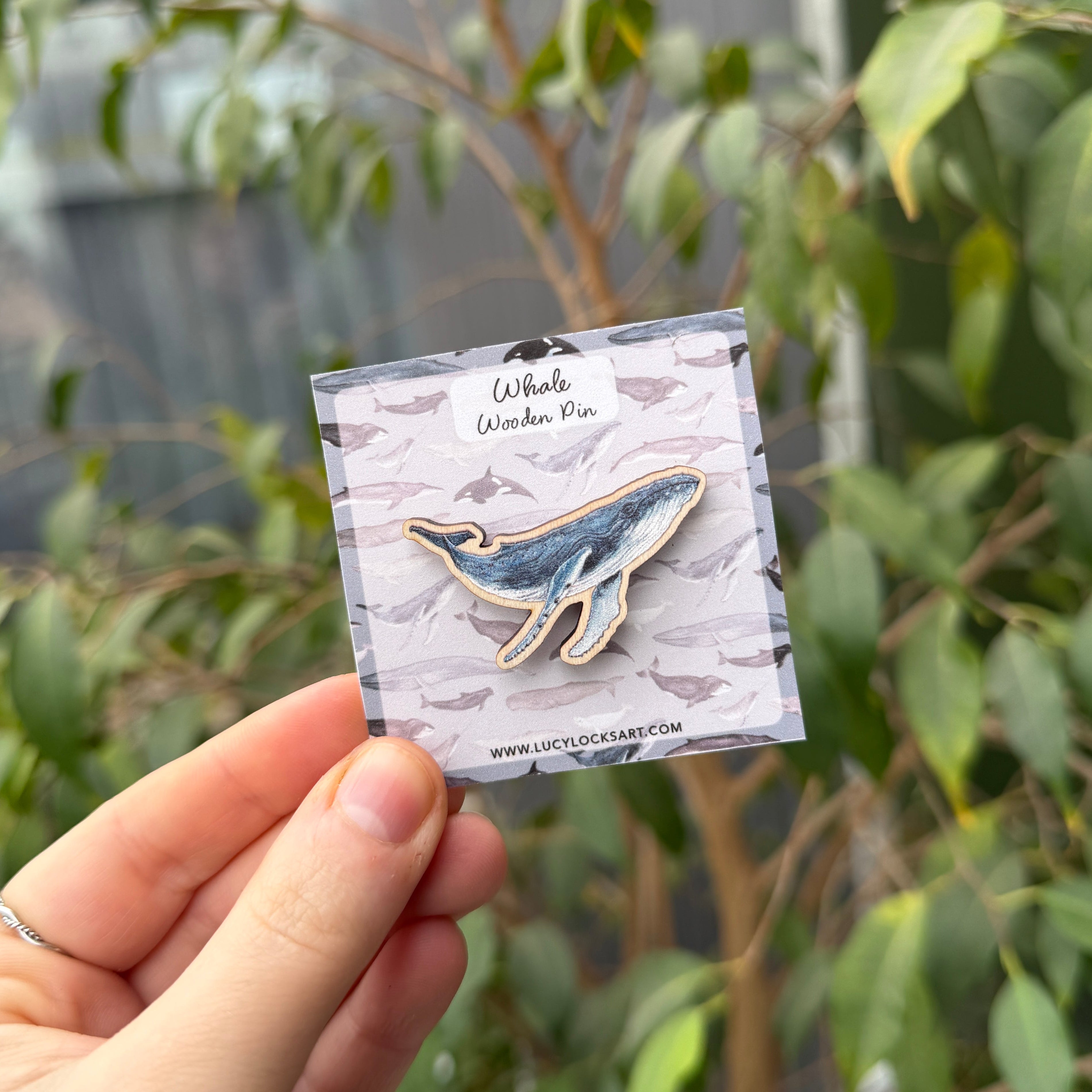 Hand holding a small card with a whale wooden pin against a green leafy background